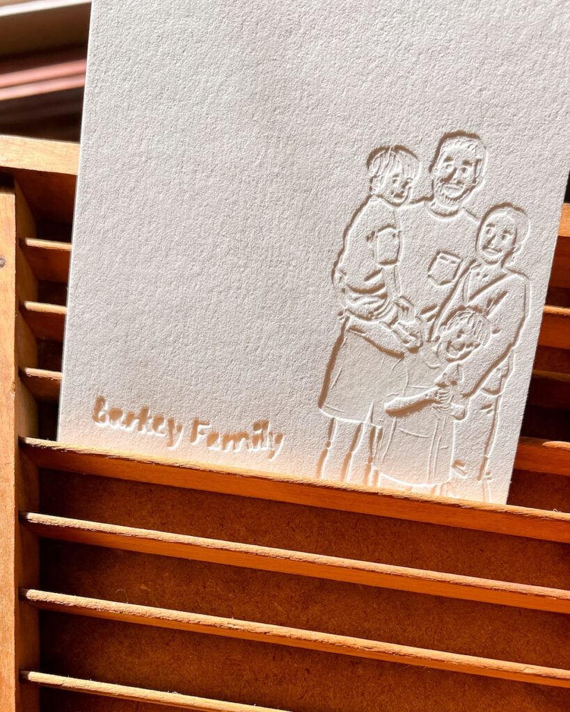 A letterpress-printed card featuring a blind "bite" impression of a family portrait. The design shows four family members (two adults and two children) standing together. The impression is visible but has no ink, emphasizing the tactile nature of the letterpress print.
