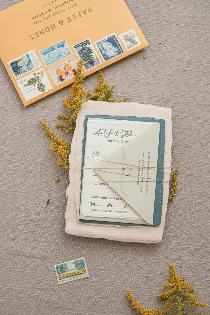 Flat lay of a whimsical wedding invitation bundle printed on handmade paper, tied with twine, and featuring illustrations of meal options. A pale yellow envelope with a full row of vintage postage stamps and elegant typography is displayed above, alongside goldenrod flowers. This image highlights charming wedding stationery and reflects consideration for 2025 USPS postage rates.