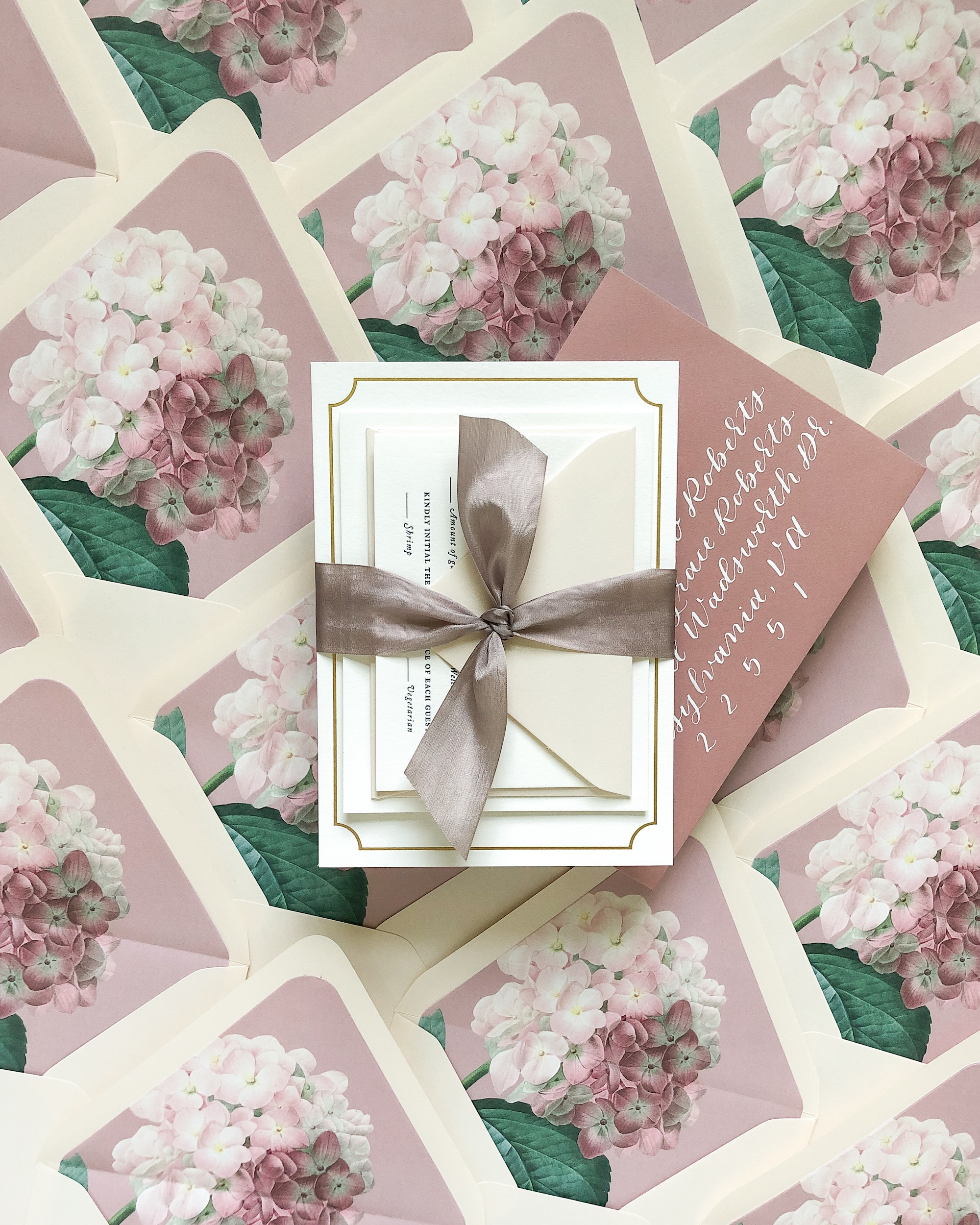 Romantic wedding invitation suite featuring cream and blush tones, elegantly tied with a taupe silk ribbon. The envelopes are hand-addressed in white calligraphy and lined with soft pink hydrangea illustrations for a cohesive floral theme. This beautifully styled flat lay highlights the thoughtful details that reflect the couple’s vision—illustrating why wedding invitations matter.