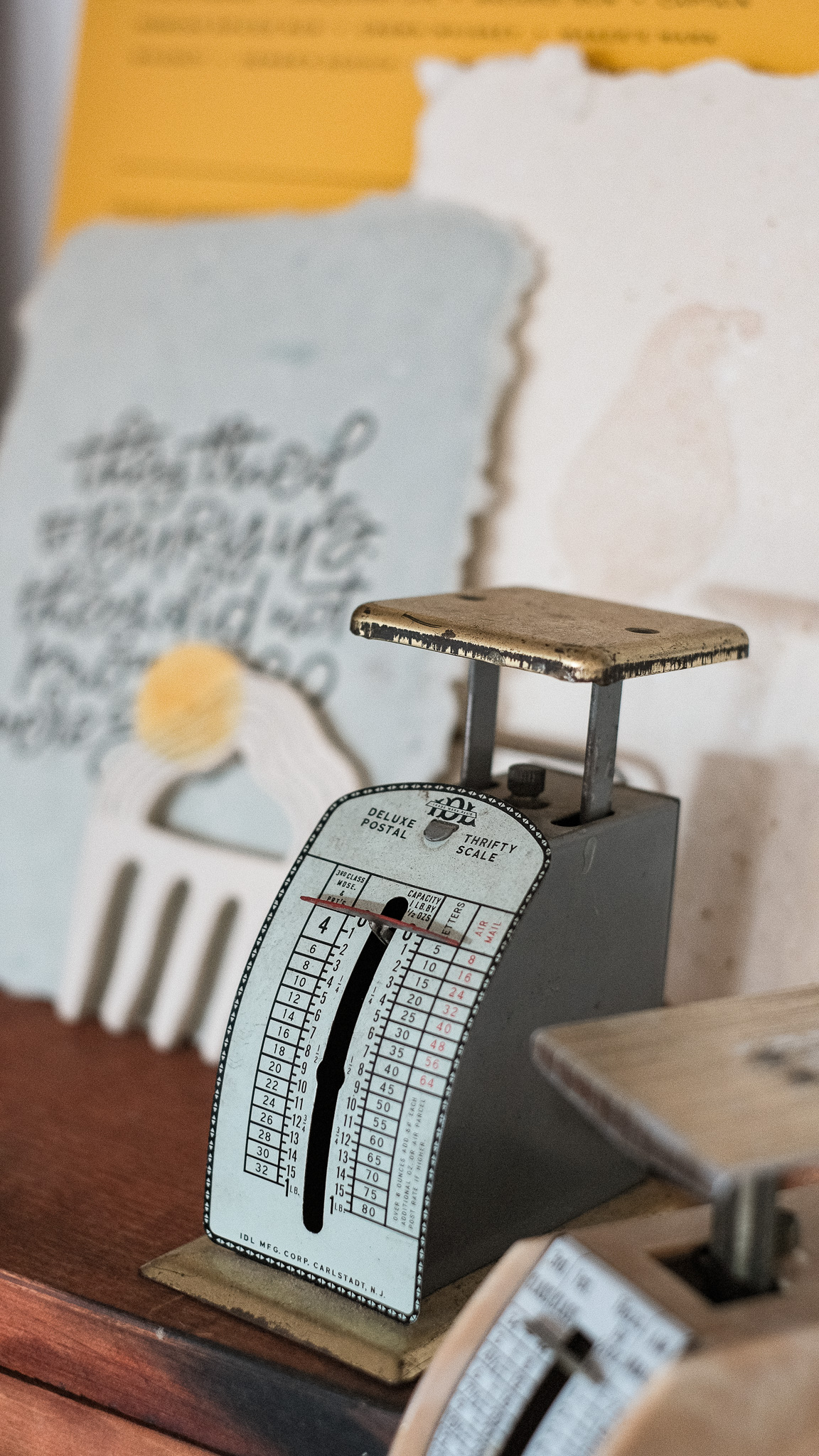 Vintage desktop postal scale labeled “Deluxe Postal Thrifty Scale” with detailed rate charts on the front, surrounded by handmade stationery and paper goods. The setup evokes a nostalgic postal theme, referencing 2025 USPS postage rates.